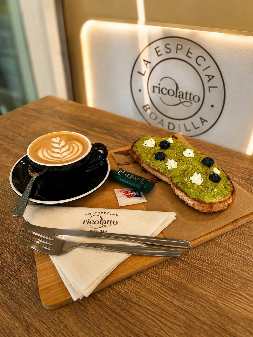 Café con leche con arte latte y la tostada La Especial sobre una mesa de madera, junto al rótulo de Ricolatto