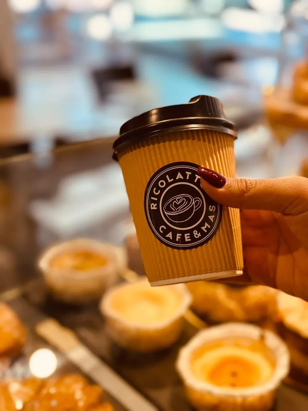 Vaso de café para llevar con el logo de Ricolatto, con la bollería de la vitrina desenfocada al fondo